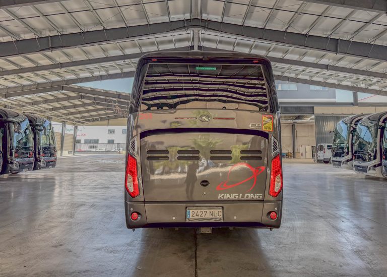 Rear view of a King Long bus inside an indoor bus depot, with other buses on both sides and a high metal ceiling above.