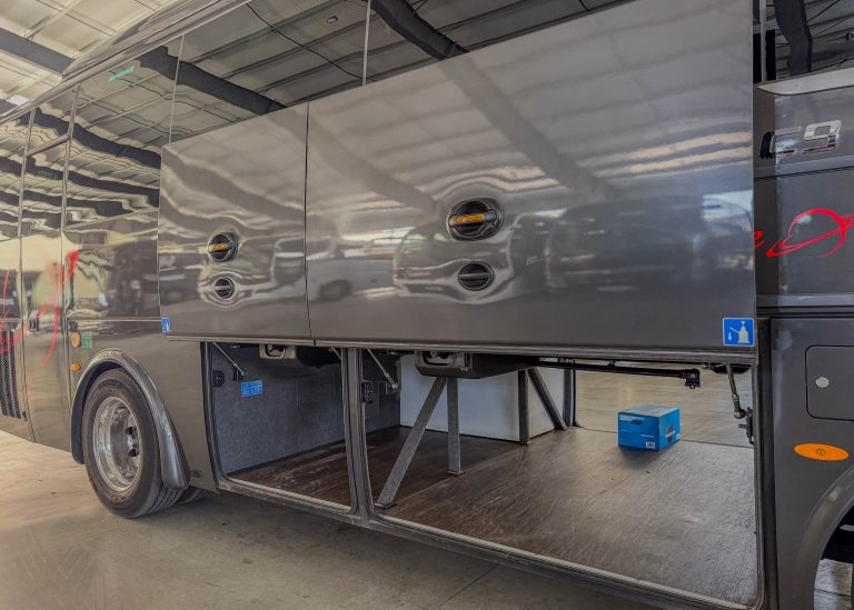Side view of a glossy black bus trailer with multiple circular storage latches and vents, reflected lights on the surface in a workshop.