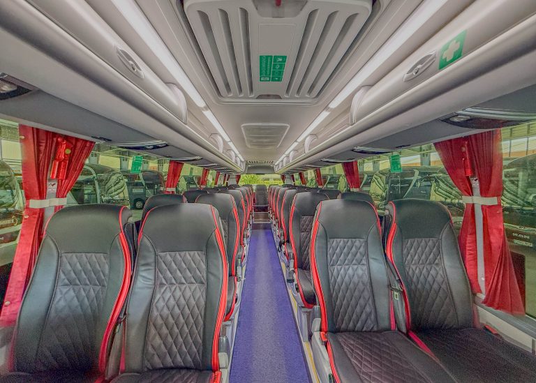 Interior of a coach bus with two columns of black seats with red piping, red curtains, and a blue aisle running down the middle.