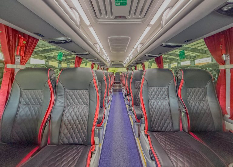 Interior of a bus with two long rows of black seats with red piping and quilted pattern, blue aisle carpet, and overhead storage.