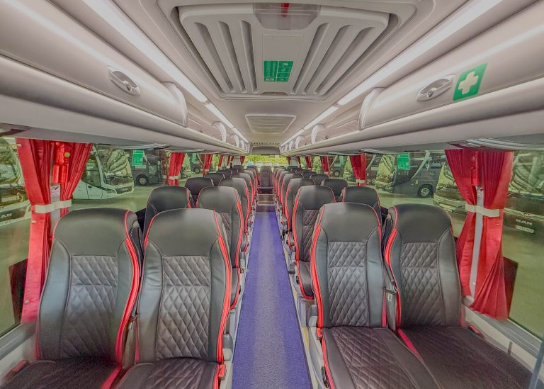 Interior of a coach bus with two columns of black seats with red piping, red curtains by the windows, and a central aisle.
