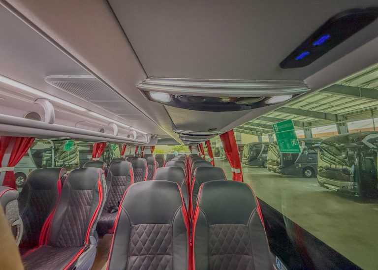 Interior of a bus with black-and-red quilted seats, red window curtains, and overhead lighting seen from the front to back.