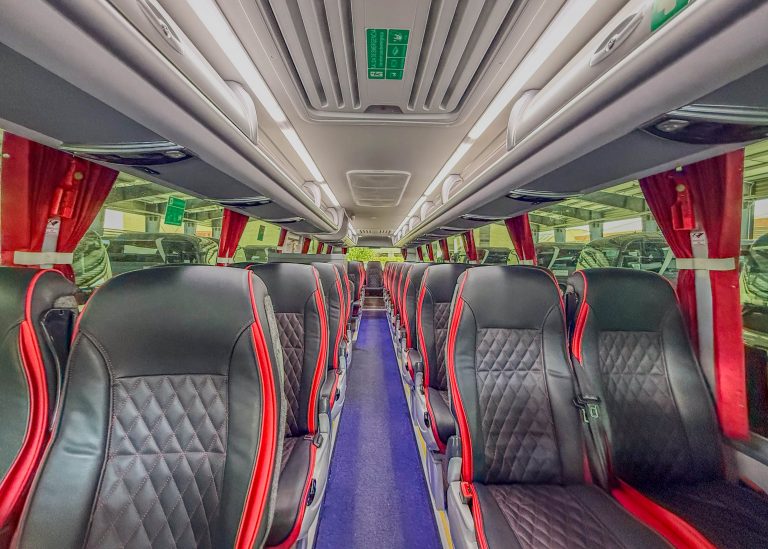 Interior of a passenger bus with rows of black seats and red piping, viewed from the aisle facing back toward the front, red curtains along windows, overhead compartments visible.