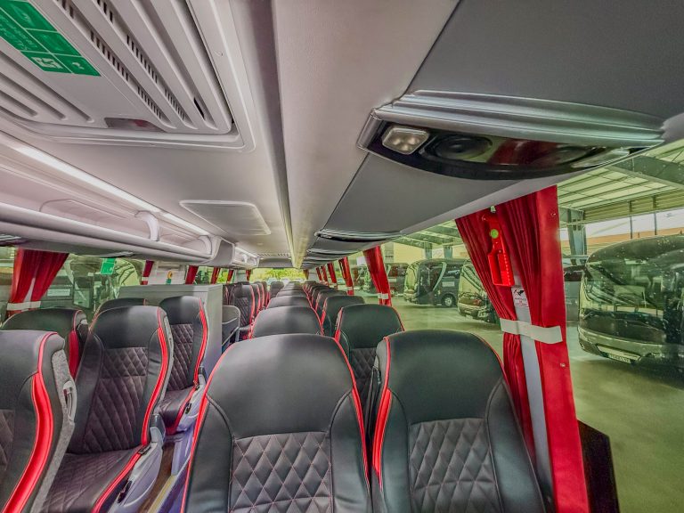 Interior of a modern tour bus with black quilted seats and red piping, red curtains, and overhead storage panels.