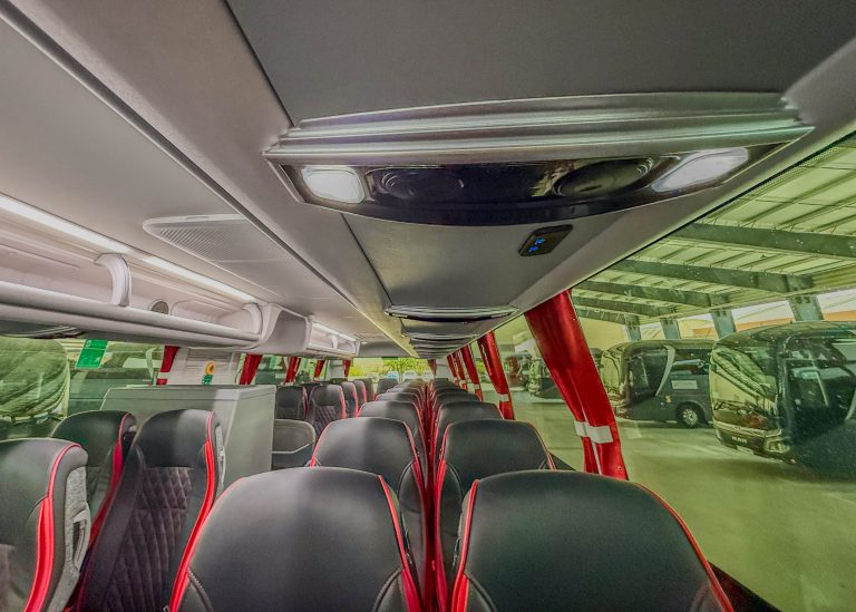 Interior of a bus with two-seat rows, black seats with red piping, and overhead luggage racks along the ceiling under bright interior lights.