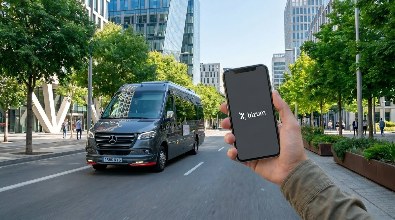Hand holding a smartphone displaying the Bizum app as a gray van drives by on a tree-lined city street in front of modern buildings.